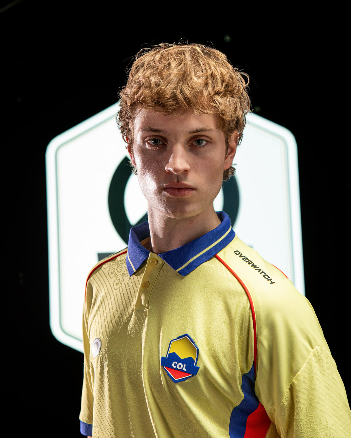 A young man with curly light brown hair wearing a yellow, blue, and red Colombia national team jersey, standing in front of a black background with a glowing white hexagonal logo.