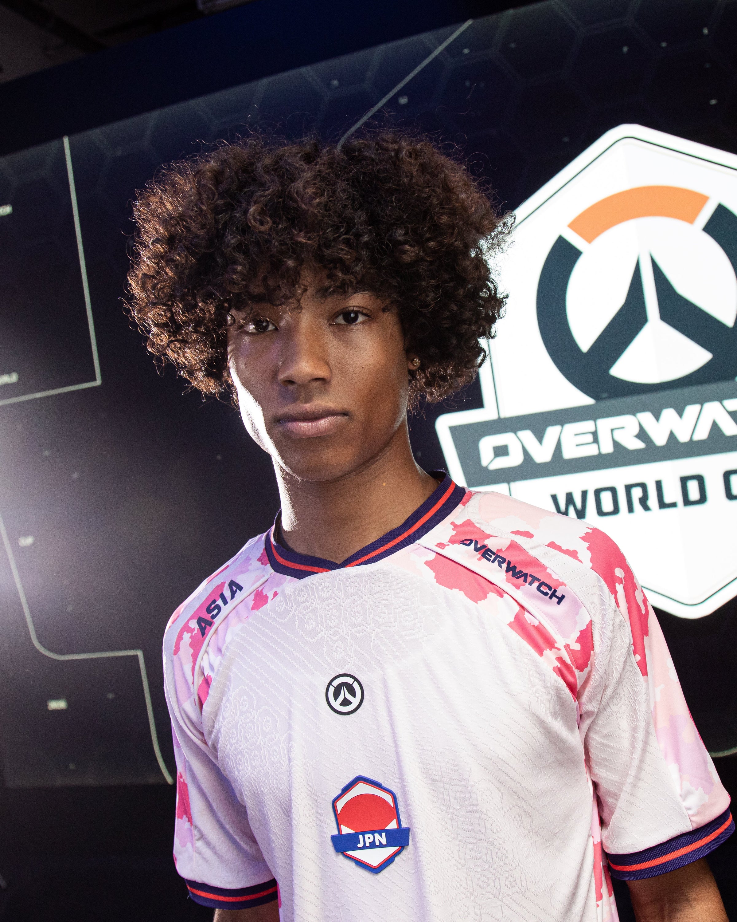 A young man with curly dark hair wearing a white and pink Overwatch World Cup jersey, standing in front of a branded Overwatch World Cup backdrop.